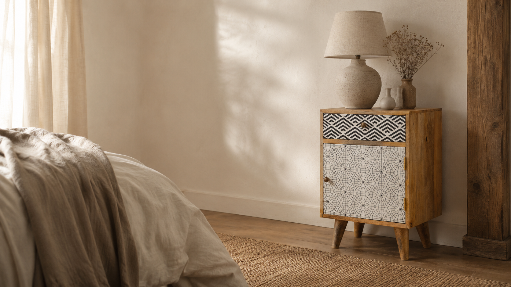 Editorial bedroom scene with a patterned wooden nightstand, linen bedding, ceramic lamp, dried stems, woven rug and soft natural daylight in a warm neutral room.