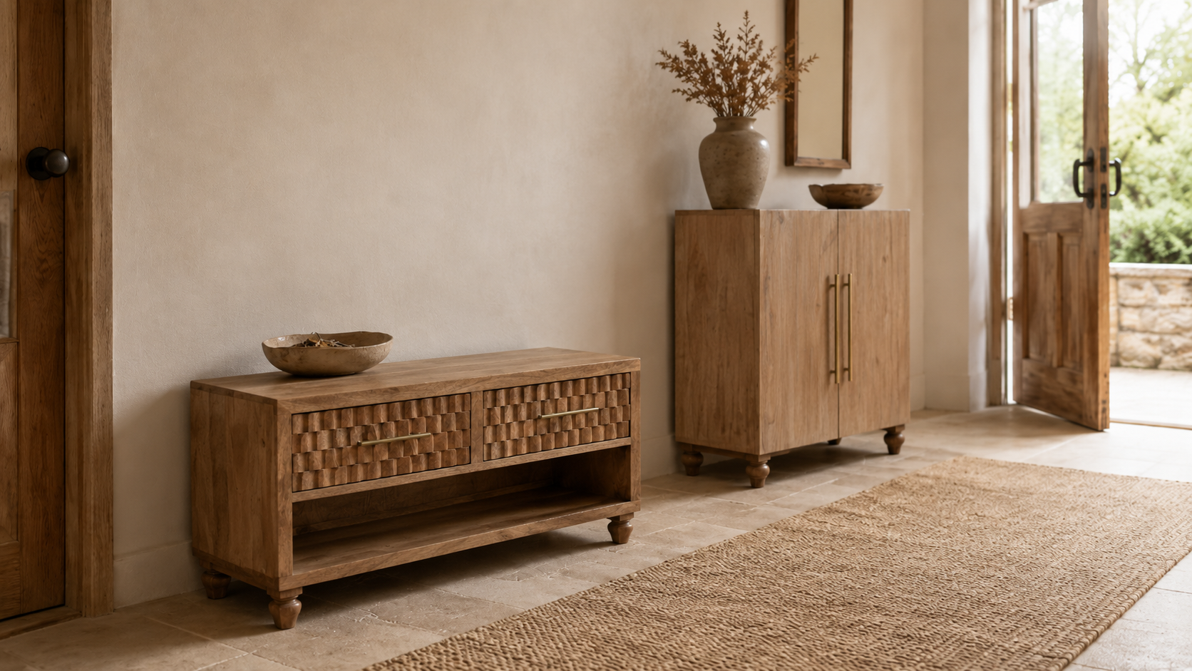 Warm rustic hallway with two wooden storage pieces, including a low carved shoe bench with open shelving and a compact two-door cabinet, styled with ceramic bowls, dried stems, a woven runner rug and soft natural light.